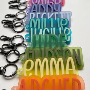 May include: A stack of colorful, translucent keychains with names printed on them. The names include: Brayton, Blake, Saigo, Addy, Beckett, Millie, Julio, Hyde, Hudson, Emma, Archer, and Brony.