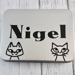 May include: A silver metal tin with a black and white cat design and the name "Nigel" printed on the front.