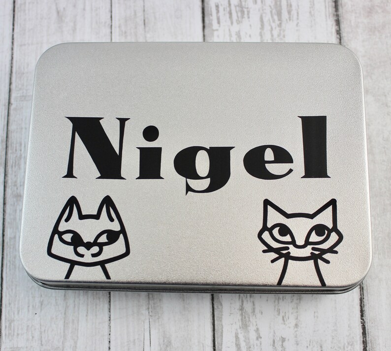 May include: A silver metal tin with a black and white cat design and the name "Nigel" printed on the front.