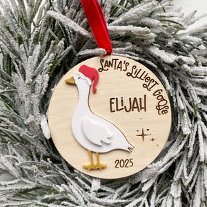 May include: A wooden Christmas ornament featuring a goose wearing a Santa hat. The ornament has the text "Santa's Lilliest Goose" and the name "Elijah" with the year "2025". A red ribbon is attached for hanging.