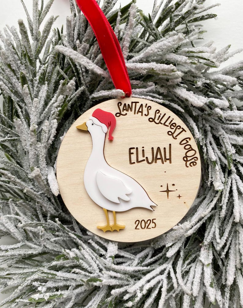 May include: A wooden Christmas ornament featuring a goose wearing a Santa hat. The ornament has the text "Santa's Lilliest Goose" and the name "Elijah" with the year "2025". A red ribbon is attached for hanging.