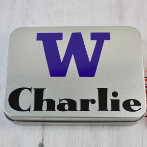 May include: A silver metal tin with a purple letter "W" and the name "Charlie" in black lettering.