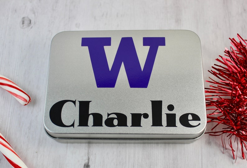 May include: A silver metal tin with a purple letter "W" and the name "Charlie" in black lettering.