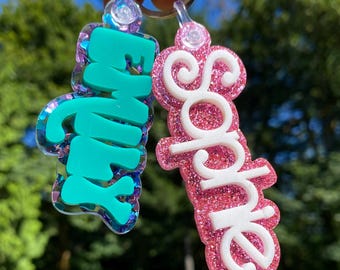 May include: Two custom keychains attached to a metal ring. One features the name Emily in teal block letters over a purple glitter base. The other features the name Sophie in white cursive letters over a pink glitter base. Both are made of acrylic.