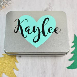 May include: A silver metal tin with a light blue heart and the name "Kaylee" written in black cursive.