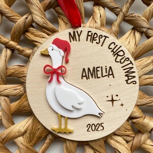 May include: A wooden Christmas ornament featuring a white goose wearing a red Santa hat and bow. The ornament is inscribed with "MY FIRST CHRISTMAS" and "AMELIA 2025". It hangs from a red ribbon and is set against a woven background.
