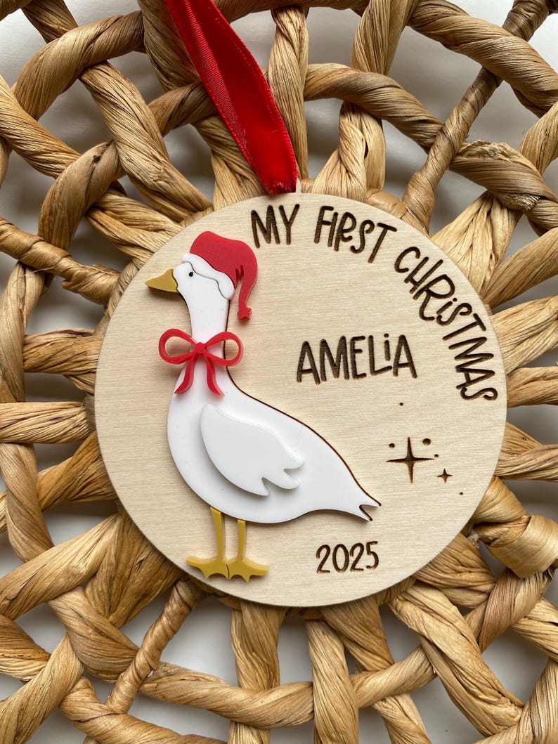 May include: A wooden Christmas ornament featuring a white goose wearing a red Santa hat and bow. The ornament is inscribed with "MY FIRST CHRISTMAS" and "AMELIA 2025". It hangs from a red ribbon and is set against a woven background.
