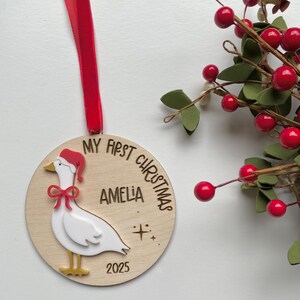 May include: A wooden Christmas ornament with a red ribbon. The ornament features a white goose wearing a Santa hat and a red bow. Text on the ornament reads "MY FIRST CHRISTMAS", "AMELIA", and "2025".