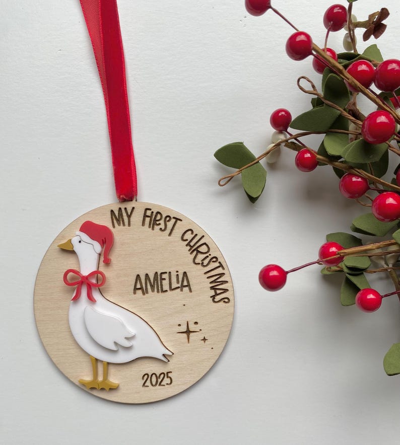 May include: A wooden Christmas ornament with a red ribbon. The ornament features a white goose wearing a Santa hat and a red bow. Text on the ornament reads "MY FIRST CHRISTMAS", "AMELIA", and "2025".