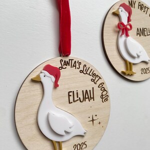 May include: Two wooden Christmas ornaments with a goose design. Each ornament features a white goose wearing a red Santa hat and gold feet. One ornament says "Santa's Lilliest Goose" and the name "Elijah" and the year "2025". The other says "My First Christmas" and the name "Amelia" and the year "2025".