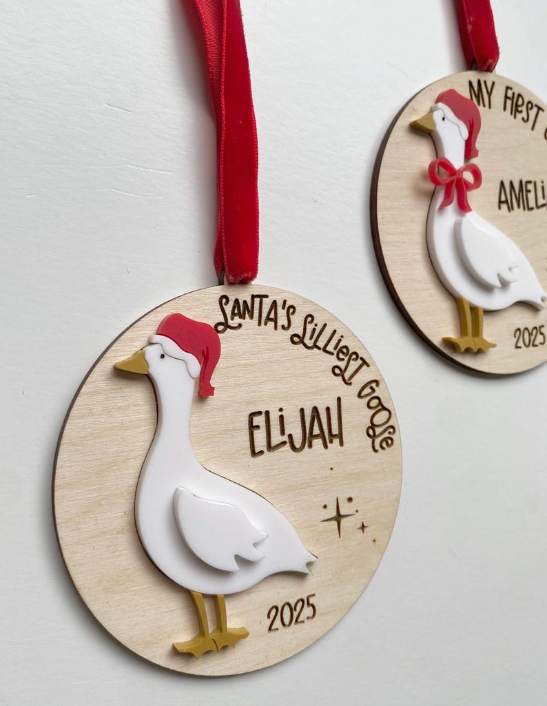 May include: Two wooden Christmas ornaments with a goose design. Each ornament features a white goose wearing a red Santa hat and gold feet. One ornament says "Santa's Lilliest Goose" and the name "Elijah" and the year "2025". The other says "My First Christmas" and the name "Amelia" and the year "2025".