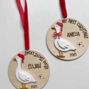 May include: Two wooden Christmas ornaments with red ribbons. Each ornament features a white goose wearing a Santa hat and the text "My First Christmas" or "Santa's Silliest Goose" with a name and the year 2025.