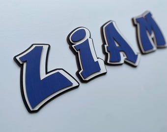 May include: Blue and white wooden letters spelling out the name "LIAM" in a graffiti style font. The letters are layered and have a 3D effect.