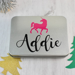 May include: A silver metal tin with a pink unicorn silhouette and the name "Addie" printed in black cursive lettering. The tin is a Christmas gift.