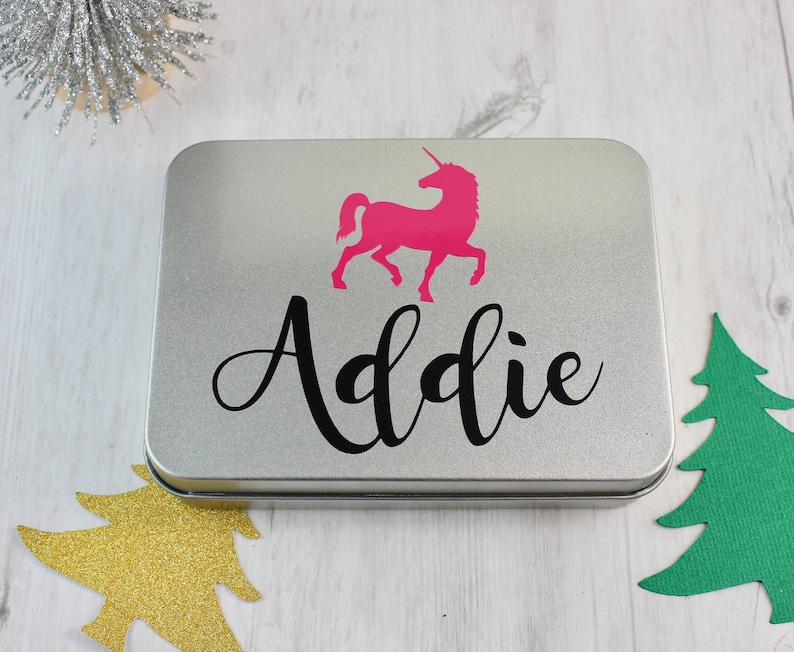 May include: A silver metal tin with a pink unicorn silhouette and the name "Addie" printed in black cursive lettering. The tin is a Christmas gift.