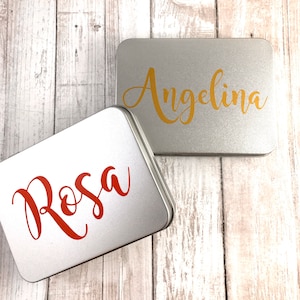 May include: Two silver metal tins with personalized names. The top tin has the name "Angelina" in gold lettering. The bottom tin has the name "Rosa" in red lettering.