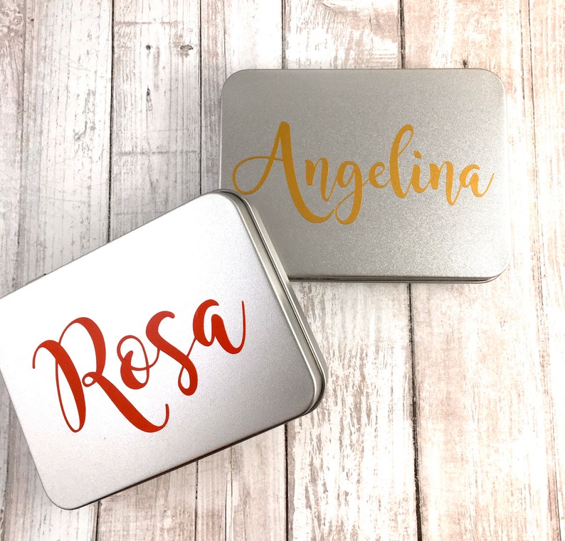 May include: Two silver metal tins with personalized names. The top tin has the name "Angelina" in gold lettering. The bottom tin has the name "Rosa" in red lettering.
