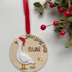 May include: A wooden Christmas ornament featuring a goose wearing a Santa hat. The ornament has the text "SANTA'S LILLIEST GOOSE" and the name "ELIJAH" engraved. A red ribbon is attached for hanging. The year "2025" is also engraved.