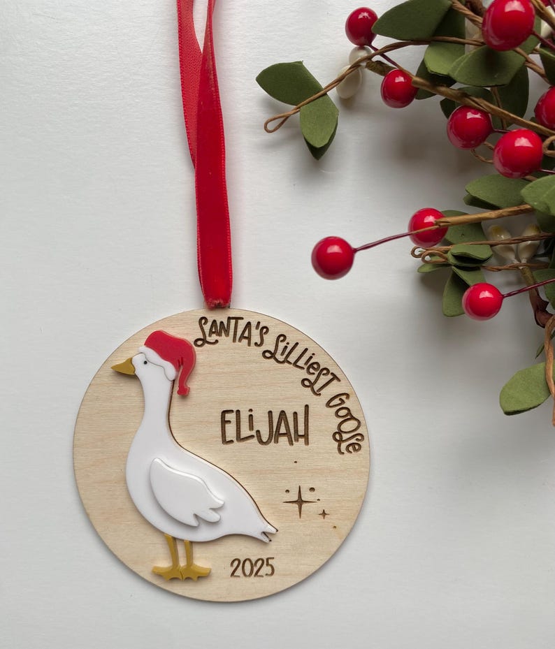 May include: A wooden Christmas ornament featuring a goose wearing a Santa hat. The ornament has the text "SANTA'S LILLIEST GOOSE" and the name "ELIJAH" engraved. A red ribbon is attached for hanging. The year "2025" is also engraved.