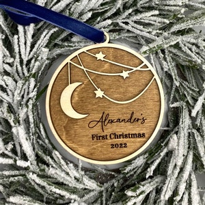 May include: A wooden Christmas ornament with a crescent moon and stars, personalized with the name "Alexander's" and the text "First Christmas 2022".