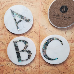 May include: Three white marble coasters with the letters A, B, and C inlaid with iridescent abalone shell. The coasters are on a brown background with a map.