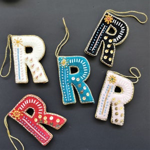 May include: Five felt letter ornaments in the shape of the letter R. Each ornament is decorated with colorful embroidery and sequins. The ornaments are in the colors white, black, teal, pink, and red.