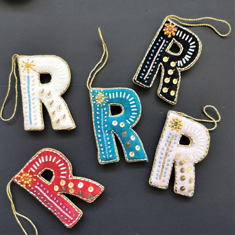 May include: Five felt letter ornaments in the shape of the letter R. Each ornament is decorated with colorful embroidery and sequins. The ornaments are in the colors white, black, teal, pink, and red.