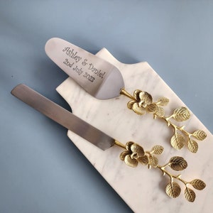 May include: A cake server and knife set with gold floral handles and a marble serving board. The server is engraved with "Ashley & Daniel 2nd July 2023". The set is designed for weddings and special occasions.