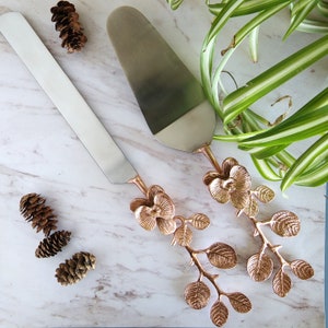 Puede incluir: Un juego de dos cuchillos para pastel de plata con mangos en forma de orqu&iacute;dea de oro rosa. Los cuchillos est&aacute;n sobre una superficie de m&aacute;rmol blanco con plantas verdes en el fondo.
