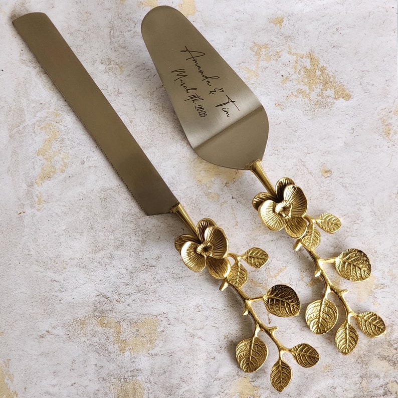 May include: A gold-colored cake knife and server set. The server is engraved with "Amanda & Tim, March 17th, 2025". Both pieces have floral and leaf accents on the handles. The set is designed for weddings and special occasions.