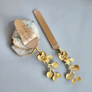 May include: A gold-colored cake server and knife set with floral handles. The server is engraved with the names "Kayleigh & Kyle" and the date "20 January 2023". The set rests on a blue and white geode slice.