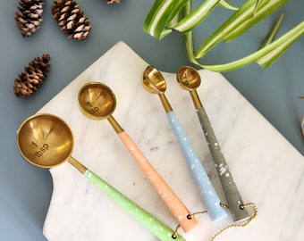 May include: Set of four gold measuring spoons with colorful speckled handles. The spoons are labeled 1/4 tsp, 1/2 tsp, 1 tsp, and 1 tbsp. The spoons are arranged on a white marble surface.