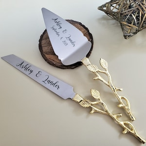 May include: A set of two silver cake servers with gold leaf branch handles. The servers are engraved with the names "Ashley & Zander" and the date "September 4, 2021".