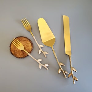 May include: Three gold-toned utensils with white twig-like handles. The utensils include a fork, a knife, and a cake server.