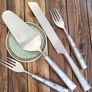 May include: A set of three silver serving utensils with mother-of-pearl handles. The utensils include a cake server, a butter knife, and a fork. The utensils are arranged on a wooden surface with a green beaded coaster.