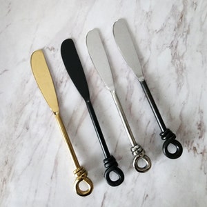 May include: Four butter knives with knotted handles. The knives are silver, gold, and black.