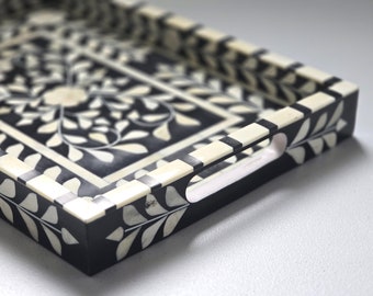 May include: Decorative black and white rectangular tray with floral and leaf inlay design. The tray has a handle on one side and is made of a dark material with a light-colored inlay. The tray's design is symmetrical and detailed.