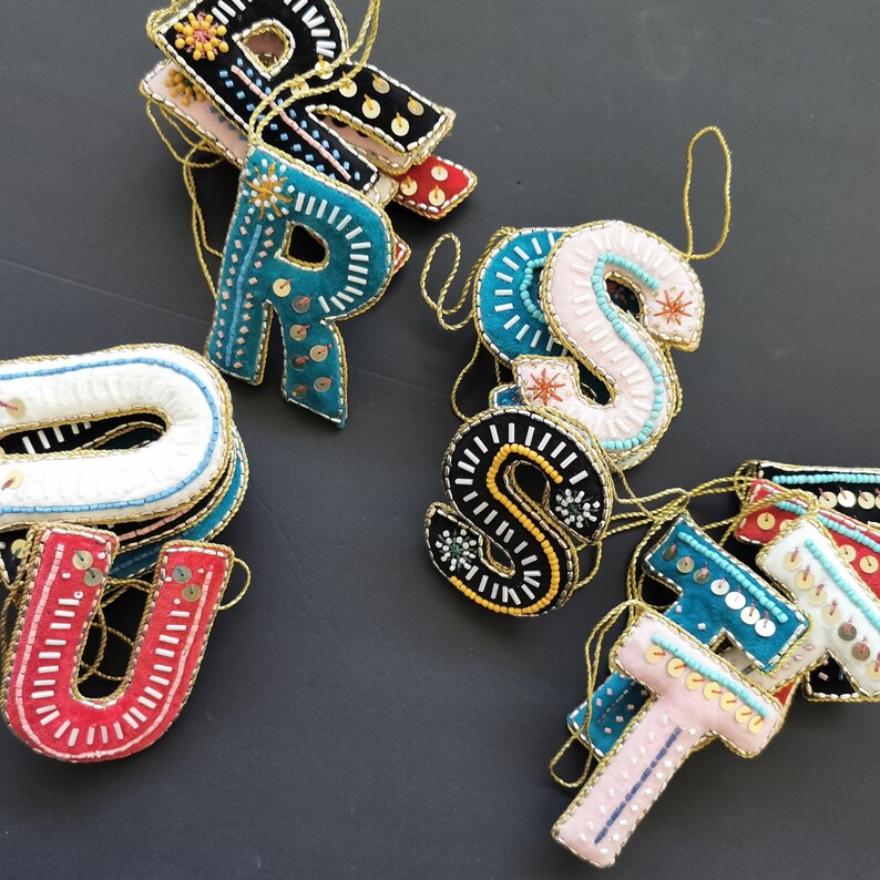 May include: A set of felt ornaments in the shape of letters, each with colorful embroidery and gold accents. The letters spell out "TRUST".