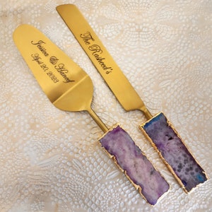 May include: Gold cake cutting set with purple agate handles. The server is engraved with "Jessica & Haneef April 20, 2023" and the knife is engraved with "The Rasheed's".