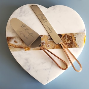 May include: A heart-shaped marble cake server with a gold trim. The server comes with two silver cake knives with copper handles. One knife is engraved with "The Knowltons Est. 8.18.2022" and the other is engraved with "Derek & Courtney".