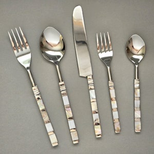 May include: A five-piece cutlery set featuring forks, spoons, and a knife. The handles are adorned with a mosaic design of white, iridescent, and silver squares. The silverware is displayed on a grey surface.