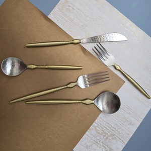 May include: A set of gold-toned cutlery with a hammered finish. The set includes a knife, fork, and two spoons.