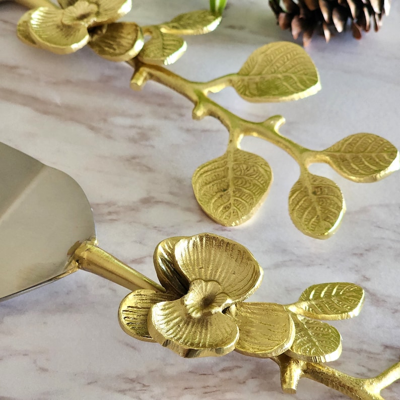 May include: A gold-colored cake server and matching server handle with floral and leaf details. The server blade is silver. The handle features a flower and leaf design, adding an elegant touch to the serving piece.