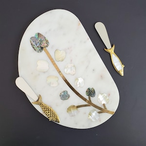 May include: White marble serving board with a gold floral design and mother-of-pearl accents. Two gold fish-shaped butter knives are included.
