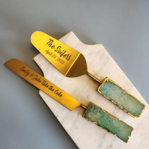 May include: Two gold-colored cake servers with engraved text. One server reads "The Soifers April 29, 2023" and the other reads "Emily & Jake Take the Cake". The handles have a gold finish and the ends have a blue-green stone with gold trim.
