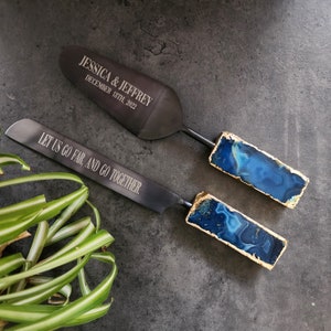 May include: A set of two black cake servers with blue agate handles. The top server is engraved with "JESSICA & JEFFREY DECEMBER 18TH, 2022". The bottom server is engraved with "LET US GO FAR, AND GO TOGETHER."