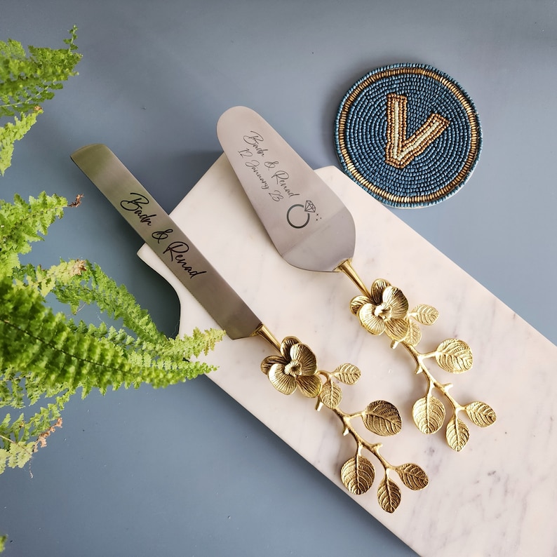 May include: A cake knife and server set with gold floral handles. The blades are engraved with "Bach & Renad" and the date "12 January 23". A blue beaded coaster with a gold "V" is also present.
