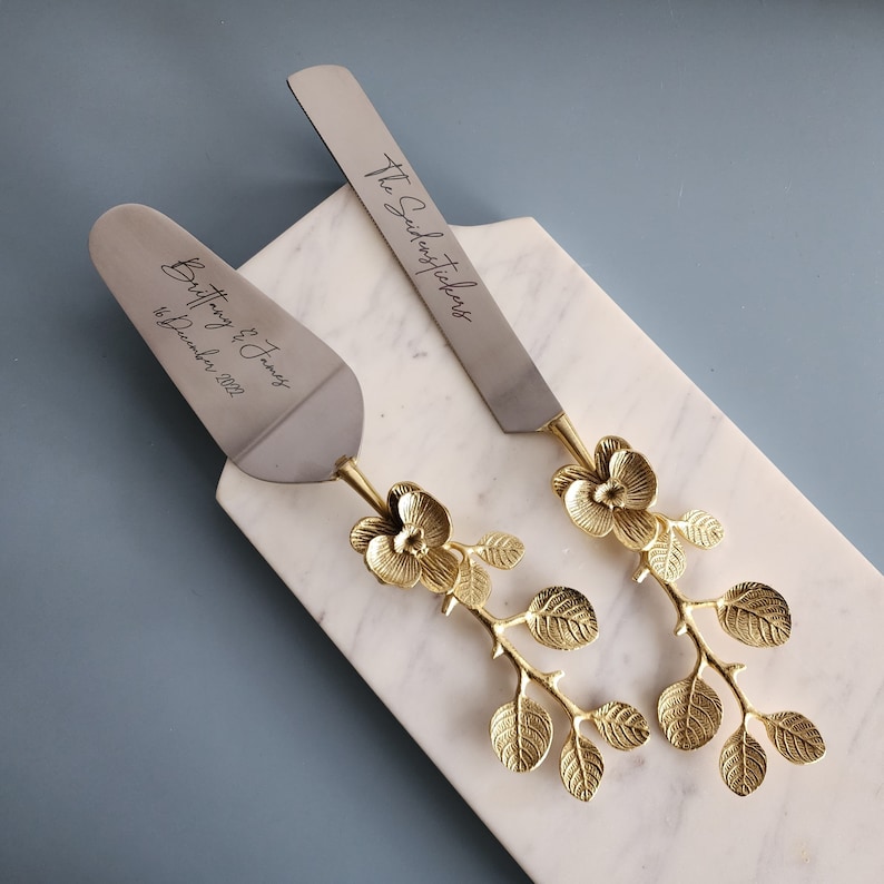 May include: A silver cake server and knife set with gold floral handles, resting on a white marble board. The server is engraved with "Brittany & James 16 December 2022", and the knife says "The SidersTickers".