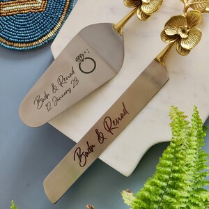 May include: A silver cake server and knife set with gold floral handles. The server has a diamond ring graphic and the text "Badr & Renad 12 January 23". The knife has the text "Badr & Renad".