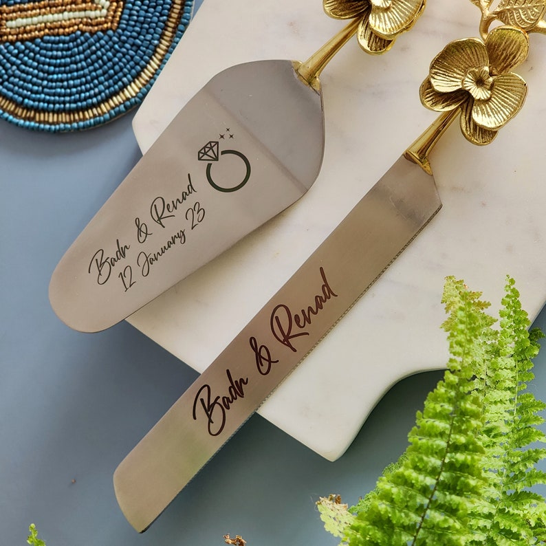 May include: A silver cake server and knife set with gold floral handles. The server has a diamond ring graphic and the text "Badr & Renad 12 January 23". The knife has the text "Badr & Renad".